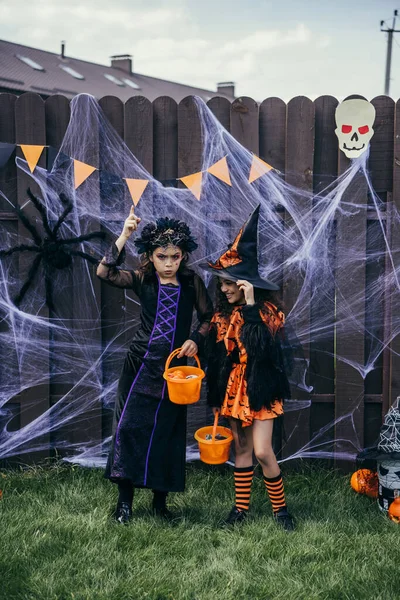 Menina em traje de Halloween segurando balde com doces perto de amigo sorridente no quintal — Fotografia de Stock