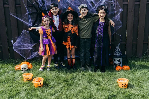 Улыбающиеся мультиэтнические дети в костюмах, смотрящие в камеру возле Хэллоуина на заборе на заднем дворе — стоковое фото