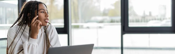 Mujer de negocios afroamericana mirando hacia otro lado cerca borrosa laptop en la oficina, pancarta - foto de stock