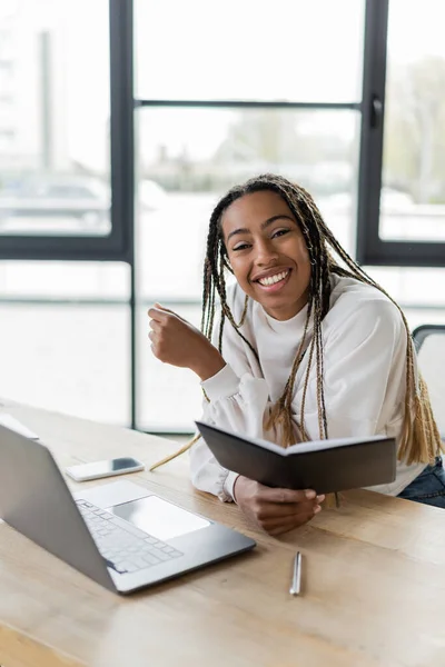Cheerful african american businesswoman holding notebook and looking at camera near devices in office — Stock Photo