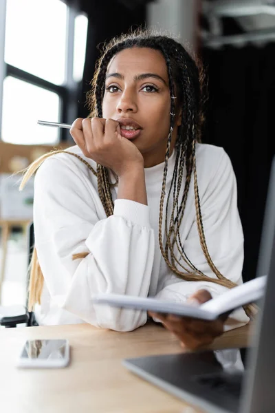 Pensiva donna d'affari africana americana che tiene penna e taccuino vicino a gadget sfocati — Foto stock