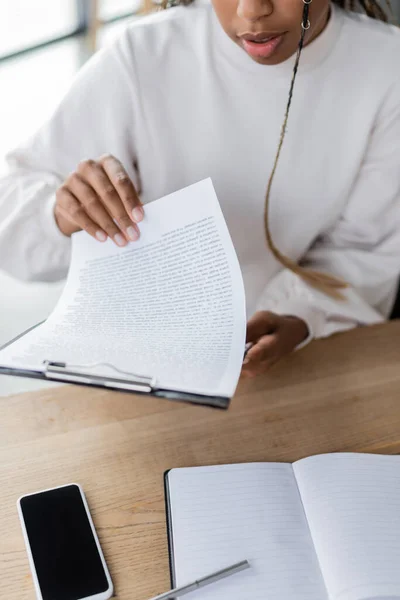 Cropped view of african american businesswoman holding clipboard near cellphone and notebook in office — Stock Photo