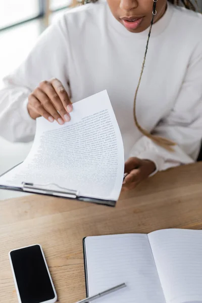 Ausschnittansicht einer afrikanisch-amerikanischen Geschäftsfrau mit Klemmbrett in der Nähe von Handy und Notizbuch im Büro — Stockfoto