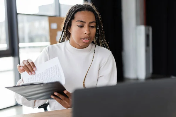 African american businesswoman holding clipboard near blurred laptop in office — Stock Photo