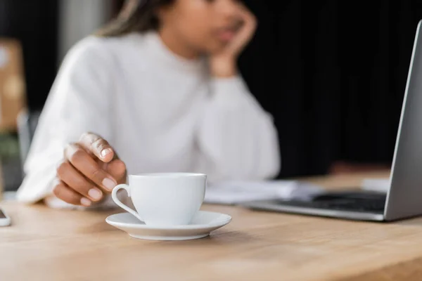 Cropped view of african american businesswoman taking cup of coffee near laptop on table — Stock Photo