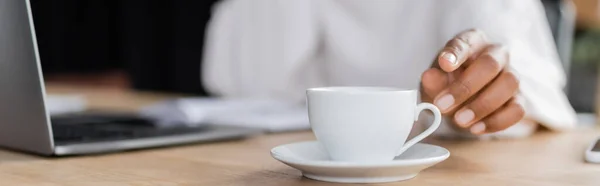 Vue recadrée d'une femme d'affaires afro-américaine floue prenant une tasse près d'un ordinateur portable sur la table au bureau, bannière — Photo de stock