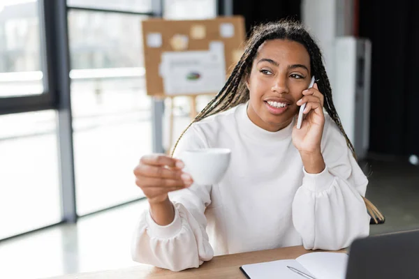 Femme d'affaires afro-américaine souriante tenant une tasse et parlant sur un smartphone au bureau — Photo de stock