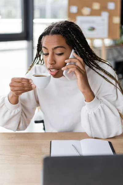 Afroamerikanische Geschäftsfrau hält eine Tasse Kaffee in der Hand, während sie im Büro in der Nähe ihres Laptops mit dem Smartphone spricht — Stockfoto