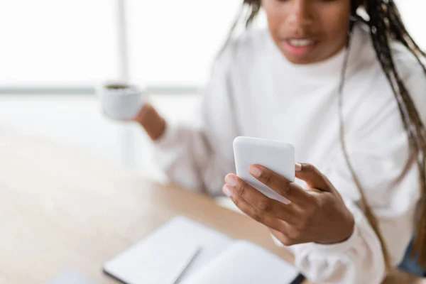 Vista recortada de la borrosa mujer de negocios afroamericana usando teléfono inteligente y taza de celebración en la oficina - foto de stock