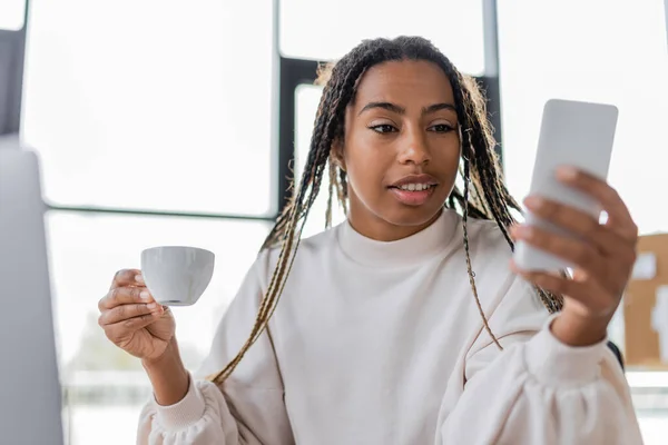 Donna d'affari afroamericana che utilizza il telefono cellulare e tiene in carica la tazza di caffè — Foto stock