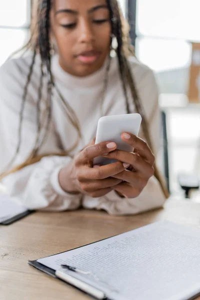 Blurred african american businesswoman using smartphone near clipboard in office — Stock Photo