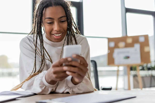 Positive african american businesswoman using blurred smartphone in office — Stock Photo