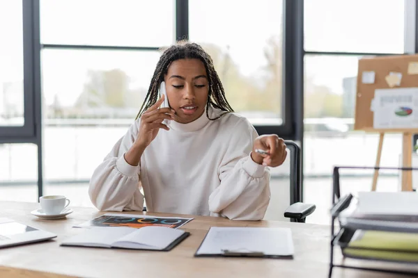 Donna d'affari afroamericana che parla al cellulare vicino a grafici e caffè in ufficio — Foto stock