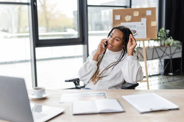 Africano americano empresária falando no smartphone perto notebook e café no escritório — Fotografia de Stock