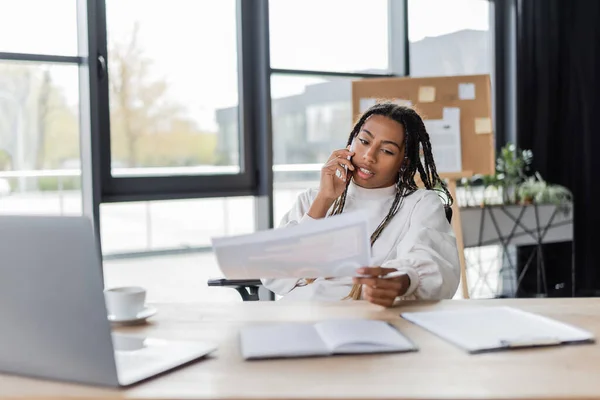 Donna d'affari afroamericana che parla su smartphone e tiene la carta vicino al laptop in ufficio — Foto stock