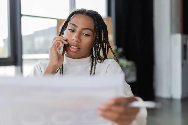 African american businesswoman holding blurred paper and talking on smartphone in office — Stock Photo