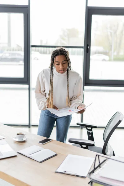 Femme d'affaires afro-américaine tenant des papiers près d'un ordinateur portable et d'un smartphone sur la table au bureau — Photo de stock