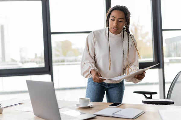 African american businesswoman holding papers near devices and cup of coffee in office — Stock Photo
