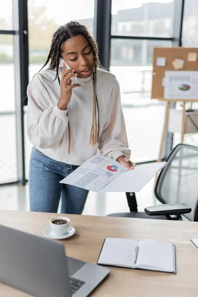 Empresaria afroamericana hablando por teléfono inteligente y sosteniendo papeles con cartas cerca del café en la oficina - foto de stock