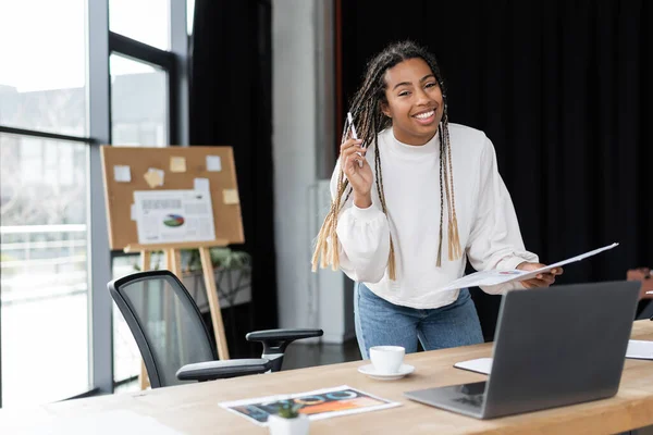 Alegre mujer de negocios afroamericana sosteniendo la carpeta de teléfonos inteligentes y papel y mirando a la cámara en la oficina - foto de stock