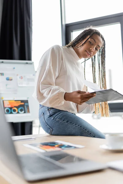 Femme d'affaires afro-américaine souriante tenant le presse-papiers et regardant l'ordinateur portable flou dans le bureau — Photo de stock