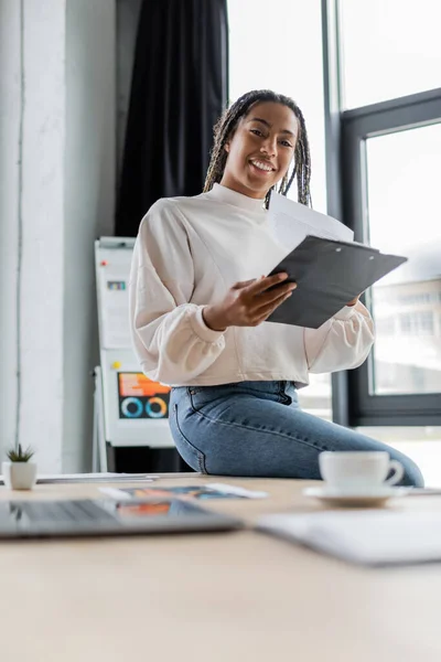 Femme d'affaires afro-américaine positive en vêtements décontractés tenant presse-papiers et regardant la caméra dans le bureau — Photo de stock