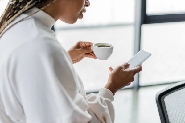 Cropped view of african american businesswoman holding cup of coffee and using smartphone in office — Stock Photo