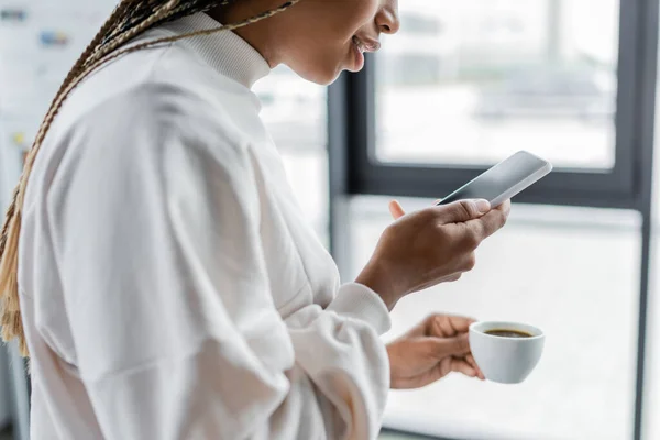 Vista cortada da empresária americana africana segurando smartphone e xícara de café no escritório — Fotografia de Stock