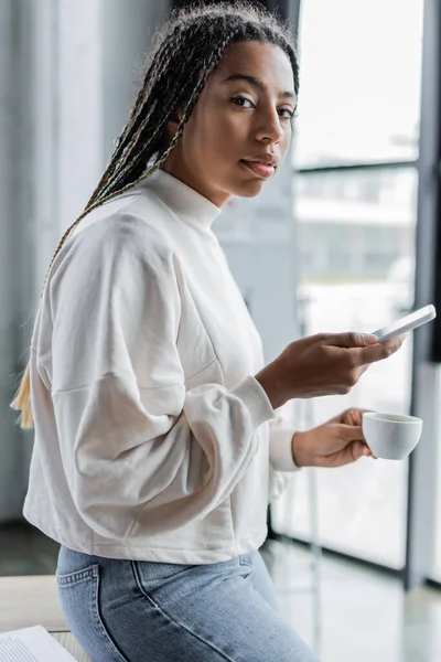 Femme d'affaires afro-américaine en sweat-shirt tenant du café et smartphone au bureau — Photo de stock