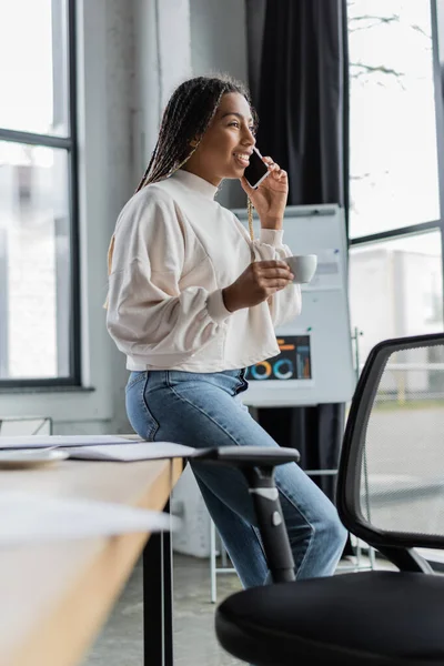 Smiling african american businesswoman in casual clothes talking on smartphone and holding cup in office — Stock Photo