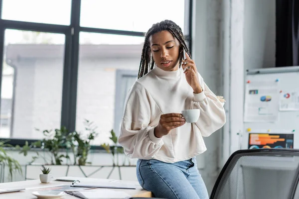 Empresaria afroamericana hablando por teléfono inteligente y sosteniendo la taza cerca del cuaderno en la mesa en la oficina — Stock Photo