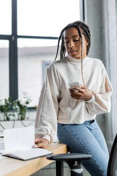 Mujer de negocios afroamericana sosteniendo un teléfono inteligente y mirando un cuaderno en la oficina - foto de stock