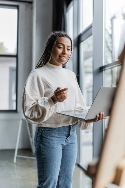 Sonriente mujer de negocios afroamericana mirando a la cámara mientras sostiene el ordenador portátil en la oficina - foto de stock