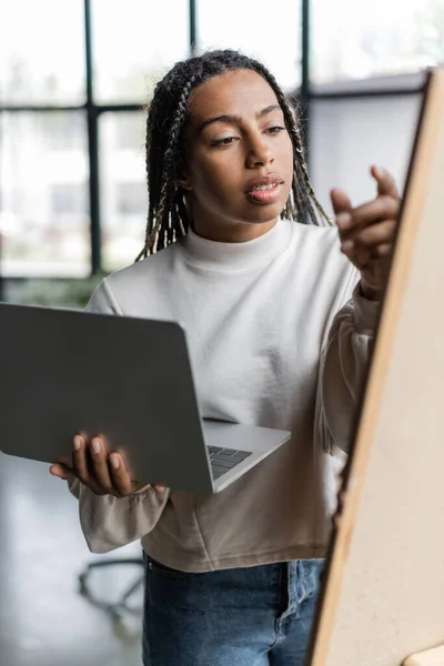 Africano americano empresária segurando laptop perto borrado bordo no escritório — Fotografia de Stock