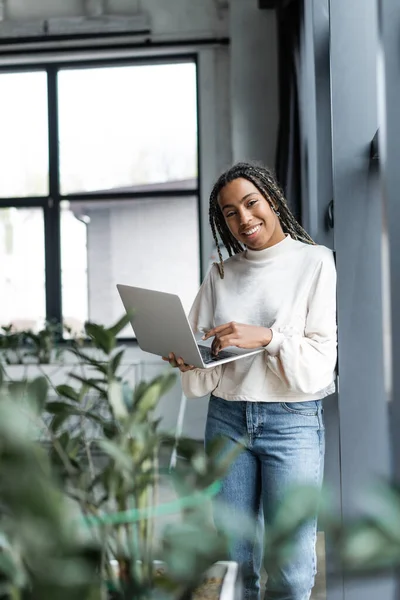 Alegre mujer de negocios afroamericana utilizando el ordenador portátil y mirando a la cámara cerca de las plantas en la oficina - foto de stock