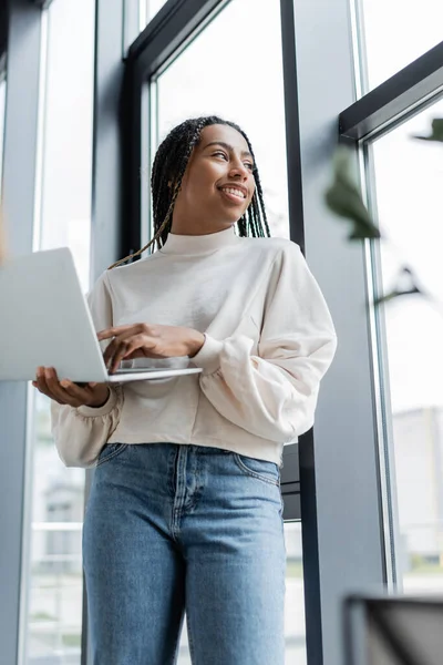 Low angle view of smiling african american businesswoman holding laptop and looking at window in office — Stock Photo