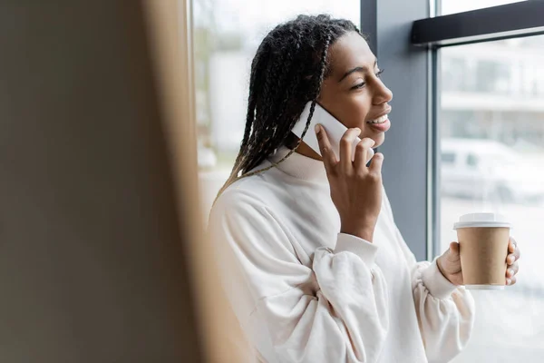 Sorridente donna d'affari afroamericana che parla su smartphone e tiene in mano il caffè per avvicinarsi alla finestra in ufficio — Foto stock