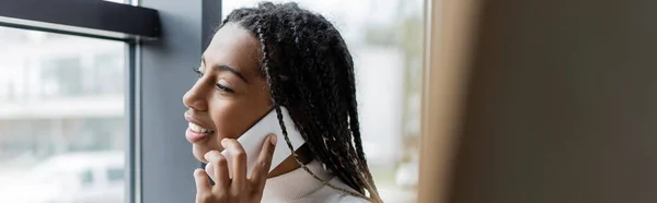 Positive african american businesswoman talking on smartphone and looking at window in office, banner — Stock Photo