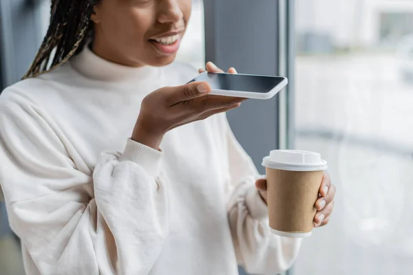 Vista recortada de la mujer de negocios afroamericana grabando mensaje de voz y sosteniendo café para ir en la oficina - foto de stock