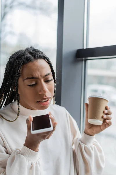Empresaria afroamericana grabando mensaje de voz en smartphone y sosteniendo café para llevar - foto de stock
