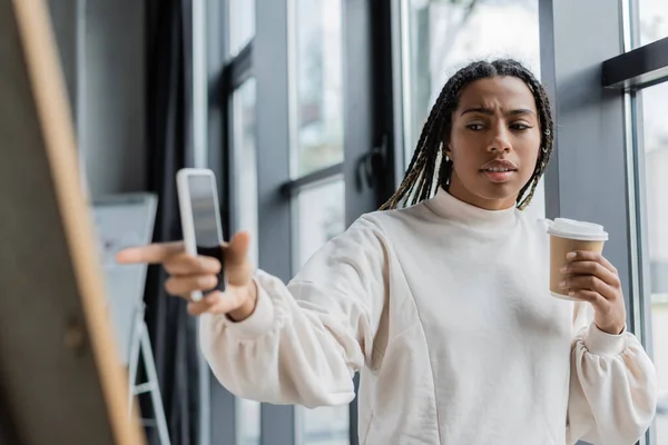 Femme d'affaires afro-américaine tenant du café pour aller et smartphone tout en pointant vers le tableau flou dans le bureau — Photo de stock