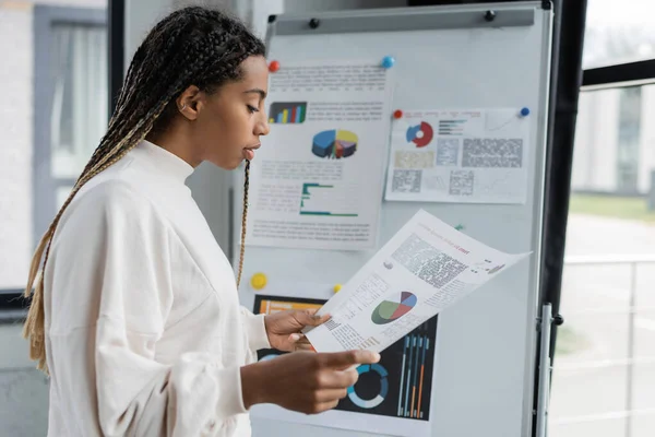 Seitenansicht einer afrikanisch-amerikanischen Geschäftsfrau, die in der Nähe eines Klappstuhls im Büro mit Diagrammen auf Papier arbeitet — Stockfoto