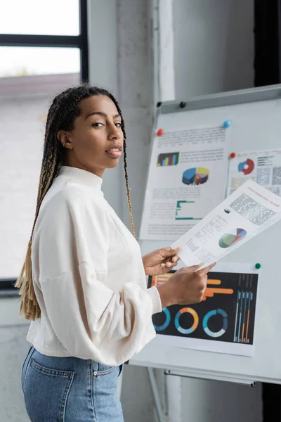 African american businesswoman holding document and looking at camera near flip chart in office — Stock Photo