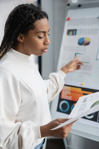 African american businesswoman holding paper and pointing at blurred flip chair in office — Stock Photo