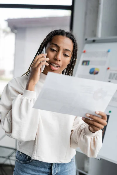 Empresaria afroamericana hablando por teléfono inteligente y sosteniendo papel en la oficina - foto de stock