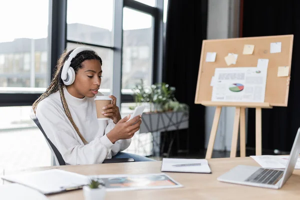 African american businesswoman in headphones holding coffee to go and using smartphone in office — Stock Photo