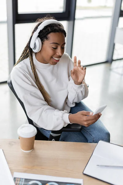 Smiling african american businesswoman in headphones having video call on smartphone in office — Stock Photo