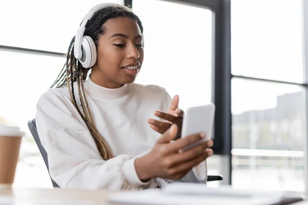 Sonriente mujer de negocios afroamericana en auriculares que tienen chat de vídeo en el teléfono inteligente cerca de café en la oficina - foto de stock