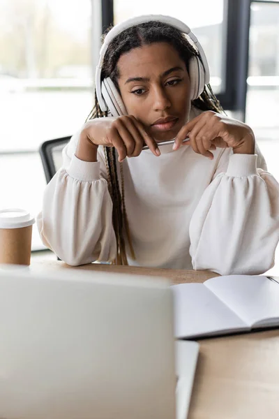 Donna d'affari afroamericana in cuffia con penna vicino al notebook e laptop sfocato in ufficio — Foto stock