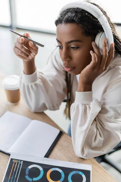 Высокий угол зрения на африканскую американскую деловую женщину в наушниках, сидящую рядом с ноутбуком и выпивку на вынос в офисе — стоковое фото